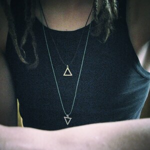 May include: A person wearing a black tank top with two silver triangle necklaces. The necklaces are layered, with the top necklace featuring a smaller triangle pendant and the bottom necklace featuring a larger triangle pendant.