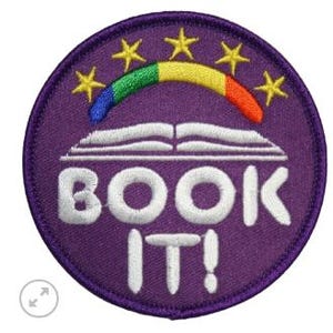 May include: A round, purple embroidered patch with the words "BOOK IT!" in white, beneath an open book design. Above the book is a rainbow arc with yellow stars. The patch has a purple border.