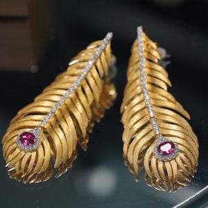 May include: A pair of gold feather-shaped earrings with a brushed matte finish. Each earring features a central row of small clear stones along the spine and a single oval-cut vibrant pink gemstone at the base, surrounded by a halo of clear stones.