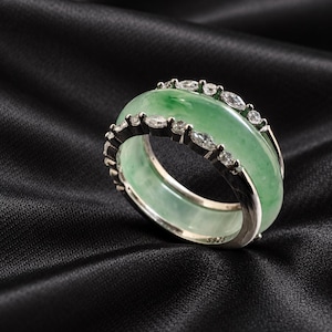 May include: A ring featuring a wide, polished green jade band set within a silver-toned metal frame. The metal structure is accented with small, clear marquise and round-cut stones along the edges. The interior band is stamped with S925.