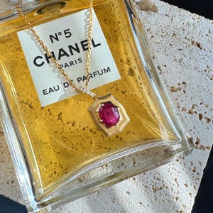 May include: A gold necklace with a pendant featuring a vibrant red gemstone. The pendant is set in a geometric frame with small, sparkling accents. The necklace is displayed near a bottle of Chanel No. 5 perfume.
