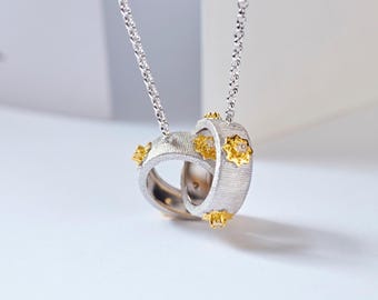 May include: Silver-toned pendant necklace featuring two interlocking rings. The rings have a textured surface and are accented with gold-colored floral designs. The necklace hangs from a delicate silver chain, creating an elegant and eye-catching piece of jewelry.