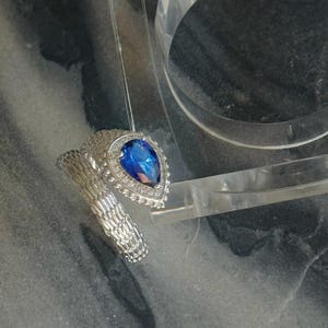 May include: A silver ring with a pear-shaped, deep blue gemstone. The ring is surrounded by smaller, clear stones and has a textured band. The ring is displayed against a marbled background.