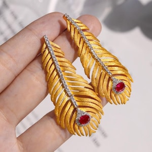 May include: A pair of gold-colored feather earrings. Each earring features a central line of small, clear stones and a red gemstone at the bottom. The earrings are designed to resemble feathers.