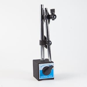 Peut inclure: Base magnétique noire et argentée avec cadran et bras réglable. La base est un cube gris foncé avec une face bleue et blanche indiquant "MAGNETIC BASE". Le bras est composé de deux tiges argentées et de joints noirs réglables.
