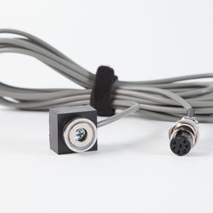 Pode incluir: Um componente elétrico preto, de forma quadrada, com um encaixe metálico circular e um cabo cinza. O cabo está enrolado e preso com uma fita de velcro preta. Um conector metálico com vários pinos é visível.