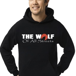 May include: Black hooded sweatshirt with the text "THE WOLF Of All Streets" in white and a red wolf silhouette. The sweatshirt has a front pocket and a drawstring hood. The person in the image is wearing the sweatshirt.