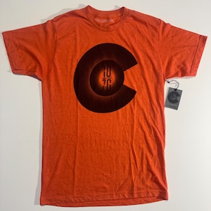 May include: Orange short-sleeved t-shirt with a large dark brown graphic of the Colorado state flag. The graphic includes the numbers "1876" in the center. A tag with a circular logo hangs from the shirt.