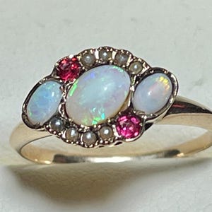 May include: A gold ring featuring a central oval opal flanked by two smaller oval opals. The setting is accented with small round pink gemstones and tiny white seed pearls arranged in a cluster around the central opal stones on a gold band.