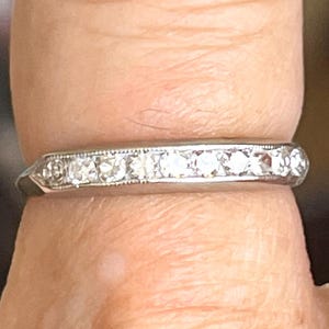 May include: A silver-coloured metal band ring featuring a row of nine small, clear, round-cut stones set in a channel. The metal setting displays a delicate milgrain texture along the edges of the stone channel, creating a refined and detailed appearance.
