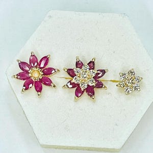 May include: Three gold-toned flower-shaped earrings. The first two earrings have a ruby-colored petal design with a central stone. The third earring is smaller, with a similar flower shape, and is adorned with clear stones.