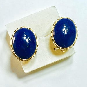 May include: A pair of gold-toned clip-on earrings featuring oval, deep blue gemstones. The gemstones are set in a gold frame with a textured design. The earrings are displayed on a white surface.