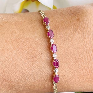 May include: A gold bracelet featuring oval-cut ruby gemstones and small, round, clear gemstones. The bracelet is worn on a wrist, with a white flower in the background. The ruby gemstones are a deep red color.