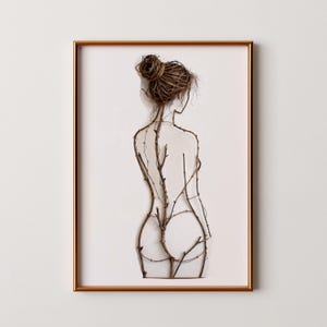 Puede incluir: Obra de arte enmarcada que presenta una figura femenina hecha de ramitas y ramas. La figura se representa de espaldas, con un moño. La obra de arte está sobre un fondo blanco y enmarcada en un marco de color cobre.