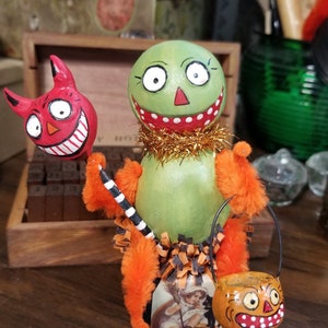 May include: A whimsical Halloween figure with a green head, orange arms, and a gold tinsel collar. It holds a red devil-shaped balloon and a jack-o'-lantern bucket. A vintage image of a child on a pumpkin is attached to the figure.