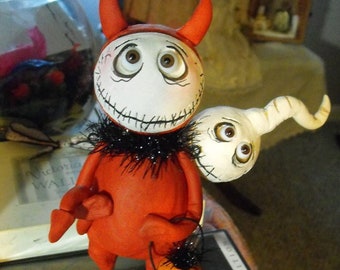 May include: A whimsical Halloween figurine of a devil-like character with a stitched smile, red body, and orange horns. It holds a pumpkin-shaped treat bag. A white ghost-like figure with two eyes is attached to the devil. The figurine is on a black base.