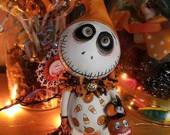 May include: A whimsical Halloween figurine with a white body, orange hat, and a stitched face. The figurine is adorned with candy corn and pumpkin details, holding a jack-o'-lantern bag and a black cat-themed broom. The figurine stands on a black base.