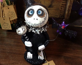 May include: A black and white Halloween figurine with a stitched face, wearing a black hat and holding a bat. The figurine is standing on a black base with glitter.