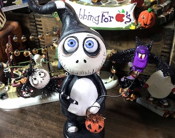 May include: A black and white figurine of a character with large blue eyes, wearing a tall hat. The figurine is holding a small orange pumpkin. Other Halloween-themed figurines are in the background.