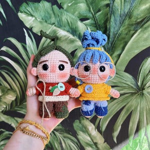 Può includere: Due bambole all'uncinetto fatte a mano. Una bambola ha una camicia rossa, pantaloni marroni e una foglia verde sulla testa. L'altra bambola ha una camicia gialla, pantaloni blu e capelli blu. Entrambe le bambole hanno bottoni e occhiali rotondi.