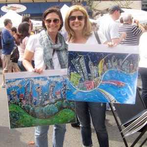 May include: Two women are holding up colorful paintings of cityscapes. The paintings are on canvas and have a whimsical style. The woman on the left is wearing a white shirt with a blue scarf and the woman on the right is wearing a blue shirt.