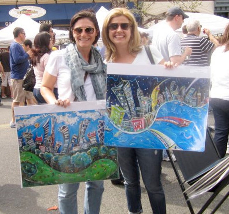 May include: Two women are holding up colorful paintings of cityscapes. The paintings are on canvas and have a whimsical style. The woman on the left is wearing a white shirt with a blue scarf and the woman on the right is wearing a blue shirt.