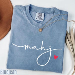 A blue t-shirt with the word "Mahj" written on it, along with a small heart symbol, is displayed on a white surface.