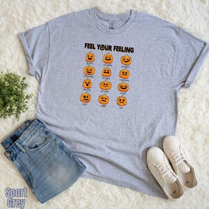 May include: A light gray t-shirt with the text "FEEL YOUR FEELING" above a grid of orange pumpkin faces, each with a different emotion. The shirt is paired with blue jeans and white sneakers.