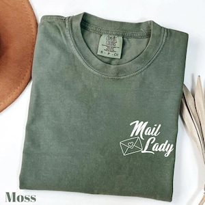 May include: Moss green t-shirt with the words "Mail Lady" in white script and an envelope graphic. The shirt is a Comfort Colors brand, size S-P-CH. A brown hat and dried plant stems are in the background.