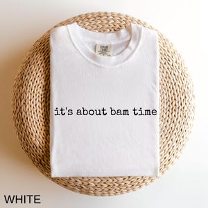 A white t-shirt with the text "it's about bam time" printed on it, laid out on a woven basket or mat.