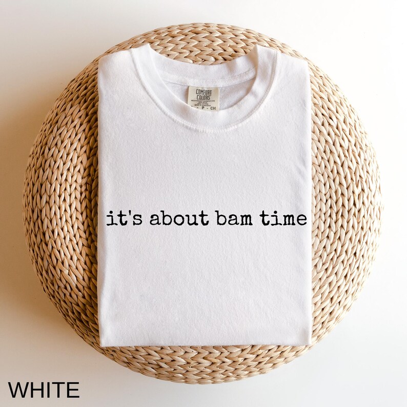 A white t-shirt with the text "it's about bam time" printed on it, laid out on a woven basket or mat.