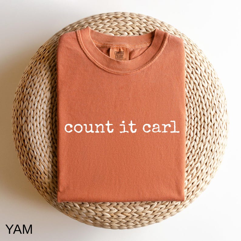 a red t-shirt with the text "count it carl" printed on it, placed on a woven basket or round seat.