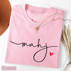 A pink t-shirt with the word "Mahj" written in cursive, accompanied by a small heart symbol, is displayed on a white background alongside a brown hat and some dried leaves.