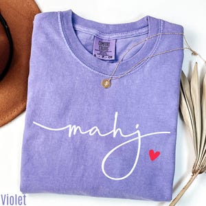 a purple t-shirt with the word "Mahj" written on it, along with a small red heart symbol. The t-shirt is folded and placed on a white surface, with a brown hat and some dried leaves visible in the background.