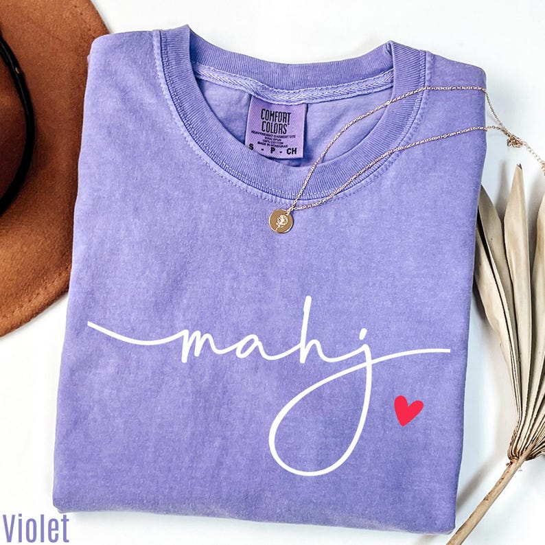 a purple t-shirt with the word "Mahj" written on it, along with a small red heart symbol. The t-shirt is folded and placed on a white surface, with a brown hat and some dried leaves visible in the background.