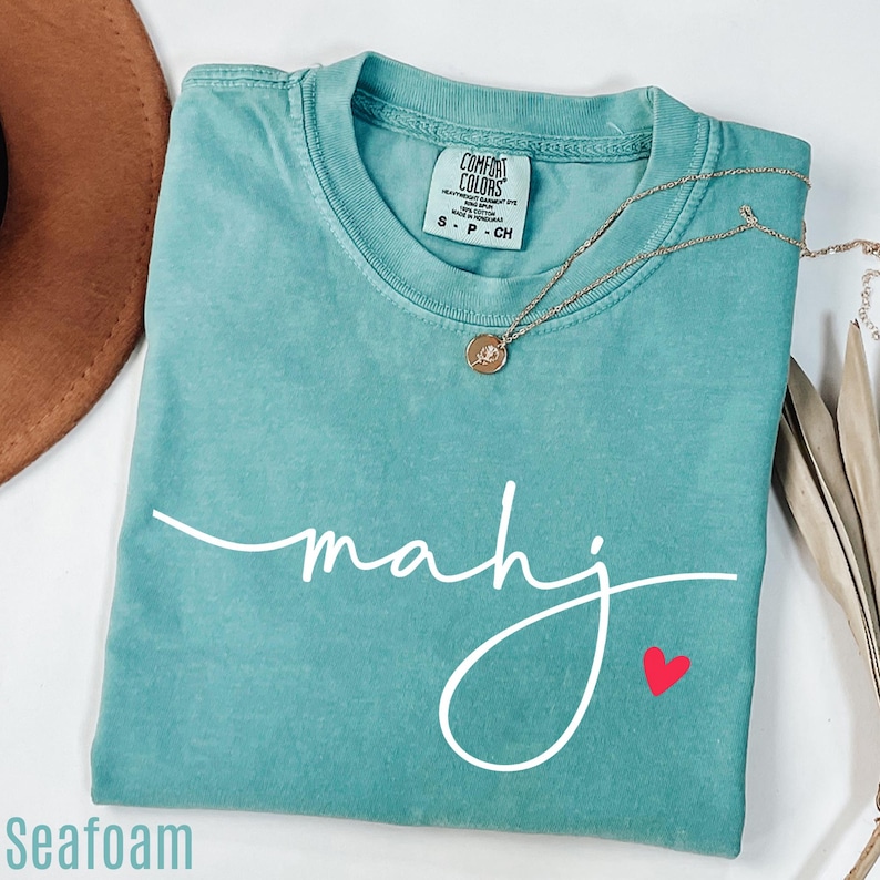 A turquoise t-shirt with the word "Mahj" written in cursive, a necklace, and a brown hat are arranged on a white surface.