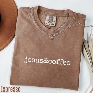 May include: Espresso-colored t-shirt with the text "jesus&coffee" in white. The shirt has a crew neck and short sleeves. A silver necklace with a small pendant is draped across the shirt. The shirt is labeled "Comfort Colors".