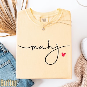a yellow t-shirt with the word "Mahj" written on it, along with a small red heart symbol. The t-shirt is placed on a white surface, and there are some jeans and a sweater visible in the background.