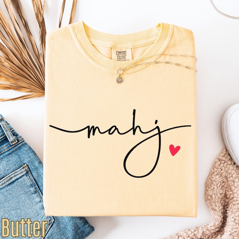 a yellow t-shirt with the word "Mahj" written on it, along with a small red heart symbol. The t-shirt is placed on a white surface, and there are some jeans and a sweater visible in the background.