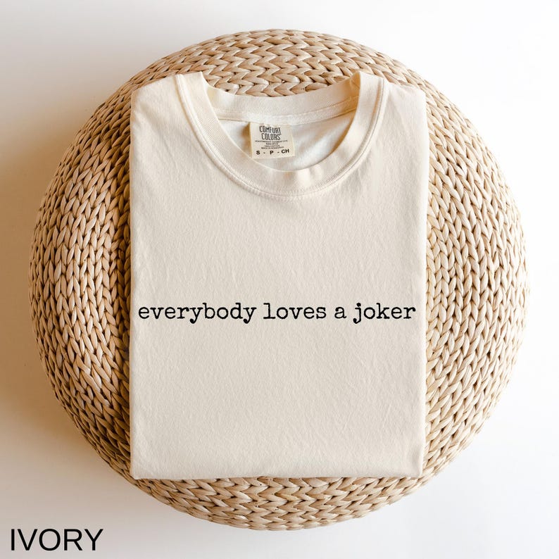 A white t-shirt with the text "Everybody loves a joker" printed on it, laid out on a woven basket or mat.