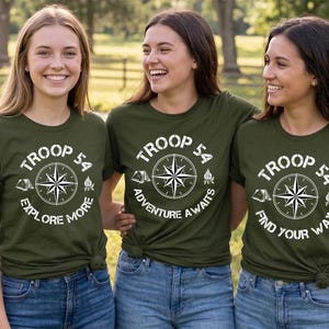 May include: Three women wearing olive green t-shirts with a white compass design and the text "TROOP 54". Each shirt has a different phrase: "EXPLORE MORE", "ADVENTURE AWAITS", and "FIND YOUR WAY". They are wearing blue jeans.