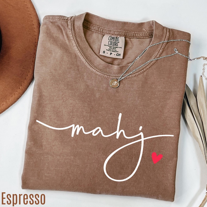 A brown t-shirt with the word "Mahj" written in cursive, a necklace, and a brown hat are arranged on a white surface.