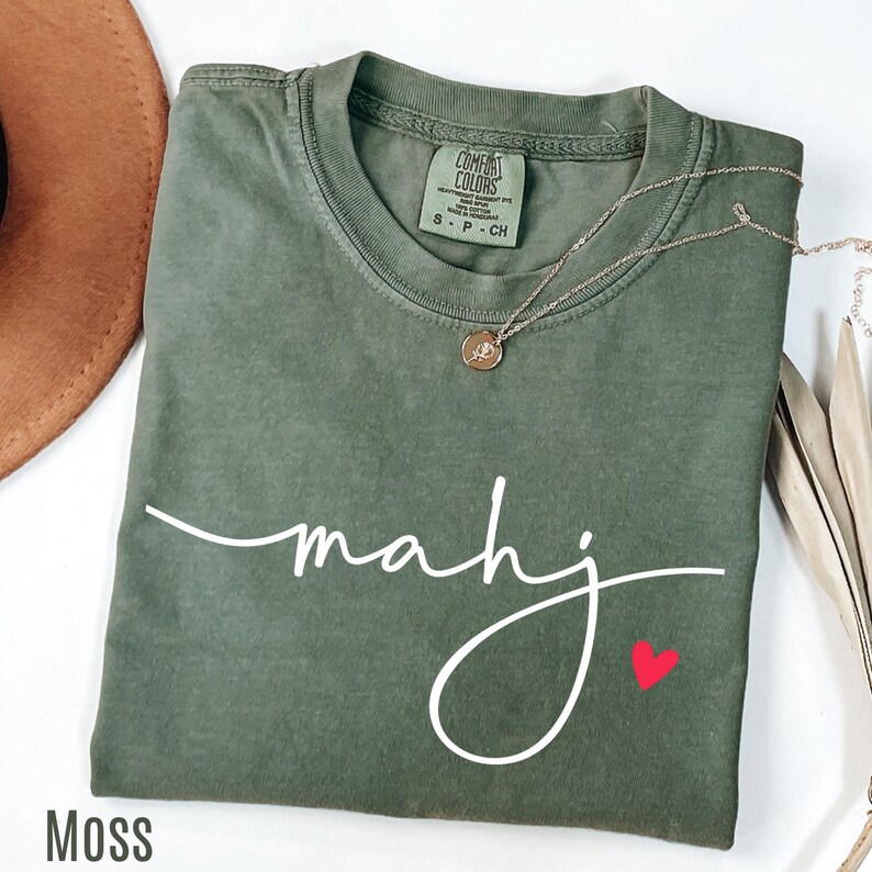 A green t-shirt with the word "mahj" written in cursive, along with a small red heart symbol, is displayed on a white surface.