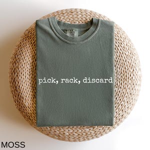 A green t-shirt with the text "pick, rack, discard" printed on it, laid out on a woven basket.