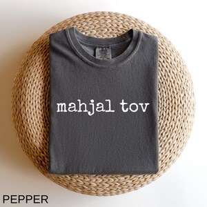 A gray t-shirt with the text "mahjal tov" printed on it, laid out on a woven basket.
