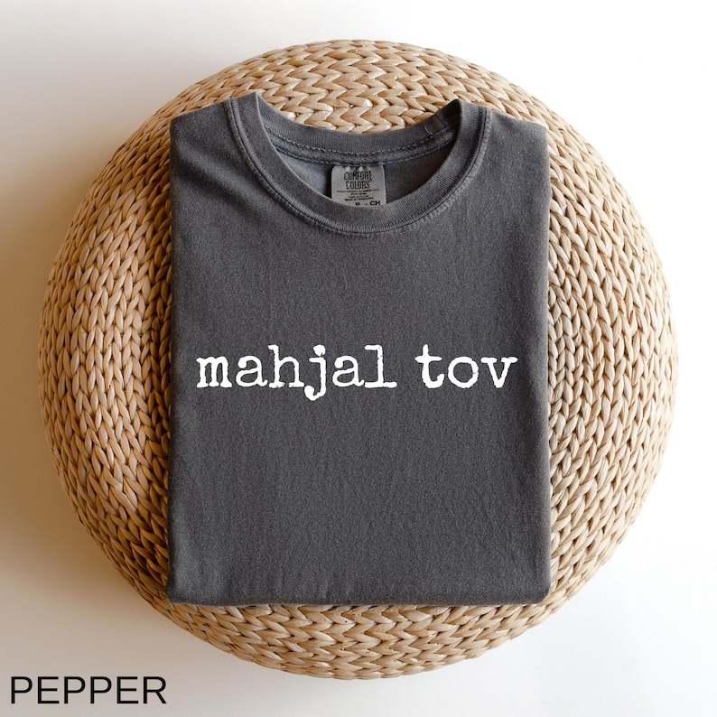 A gray t-shirt with the text "mahjal tov" printed on it, laid out on a woven basket.