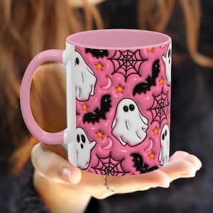 Pink 11oz ceramic mug with pink handle and interior, featuring a playful Halloween design with white ghosts, black bats, spider webs, crescent moons, and orange stars