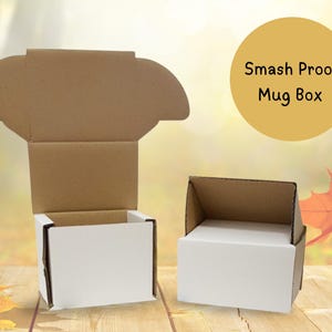 May include: Two white and brown cardboard mug boxes, one open and one closed, are displayed on a wooden surface. The boxes have a simple design with the text "Smash Proof Mug Box" in a yellow circle.