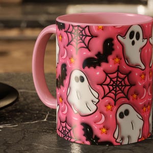 May include: Pink ceramic mug with a pink handle and interior. The mug features a repeating pattern of white cartoon ghosts, black bats, spiderwebs, stars, and crescent moons on a pink background. A Halloween-themed design.