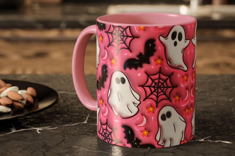 May include: Pink ceramic mug with a pink handle and interior. The mug features a repeating pattern of white cartoon ghosts, black bats, spiderwebs, stars, and crescent moons on a pink background. A Halloween-themed design.